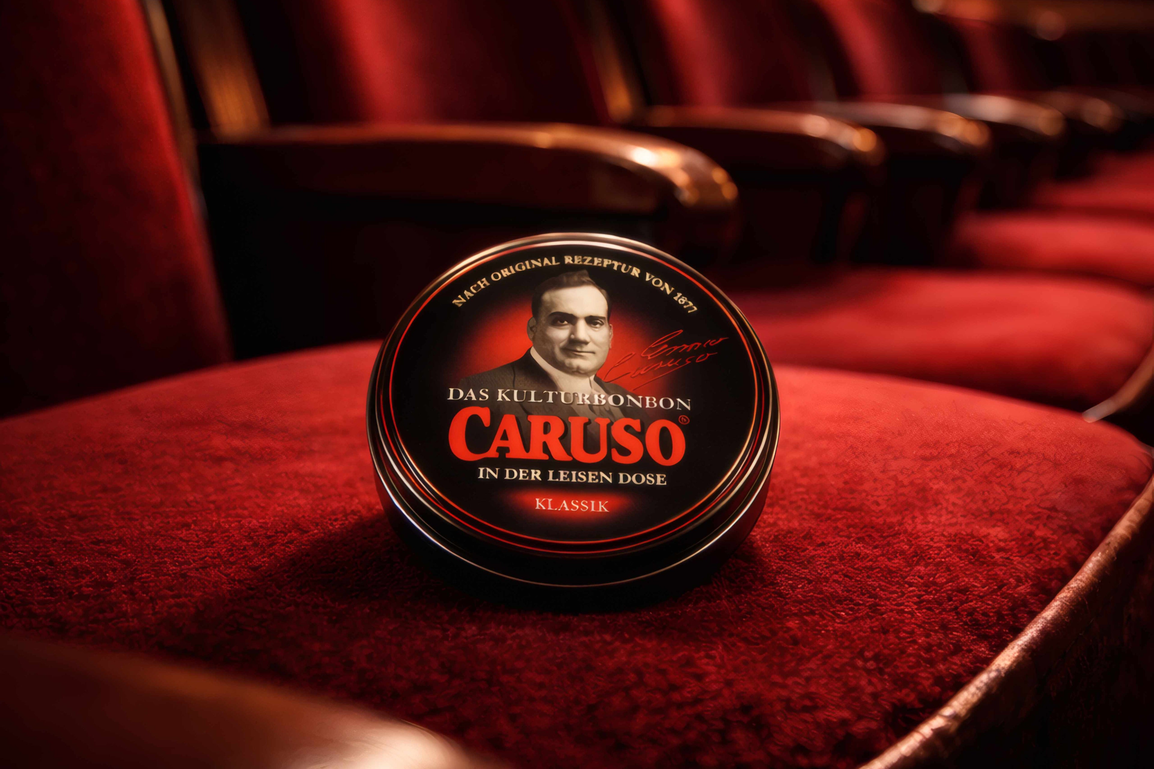 Schwarze CARUSO „Kulturbonbon“ Dose (Klassik) mit roter Beschriftung und Porträt von Enrico Caruso auf einem roten Theatersitz. Im Hintergrund sind weitere rote Samtsessel und warmes, goldenes Bühnenlicht unscharf zu erkennen – stimmungsvolle Theateratmosphäre.